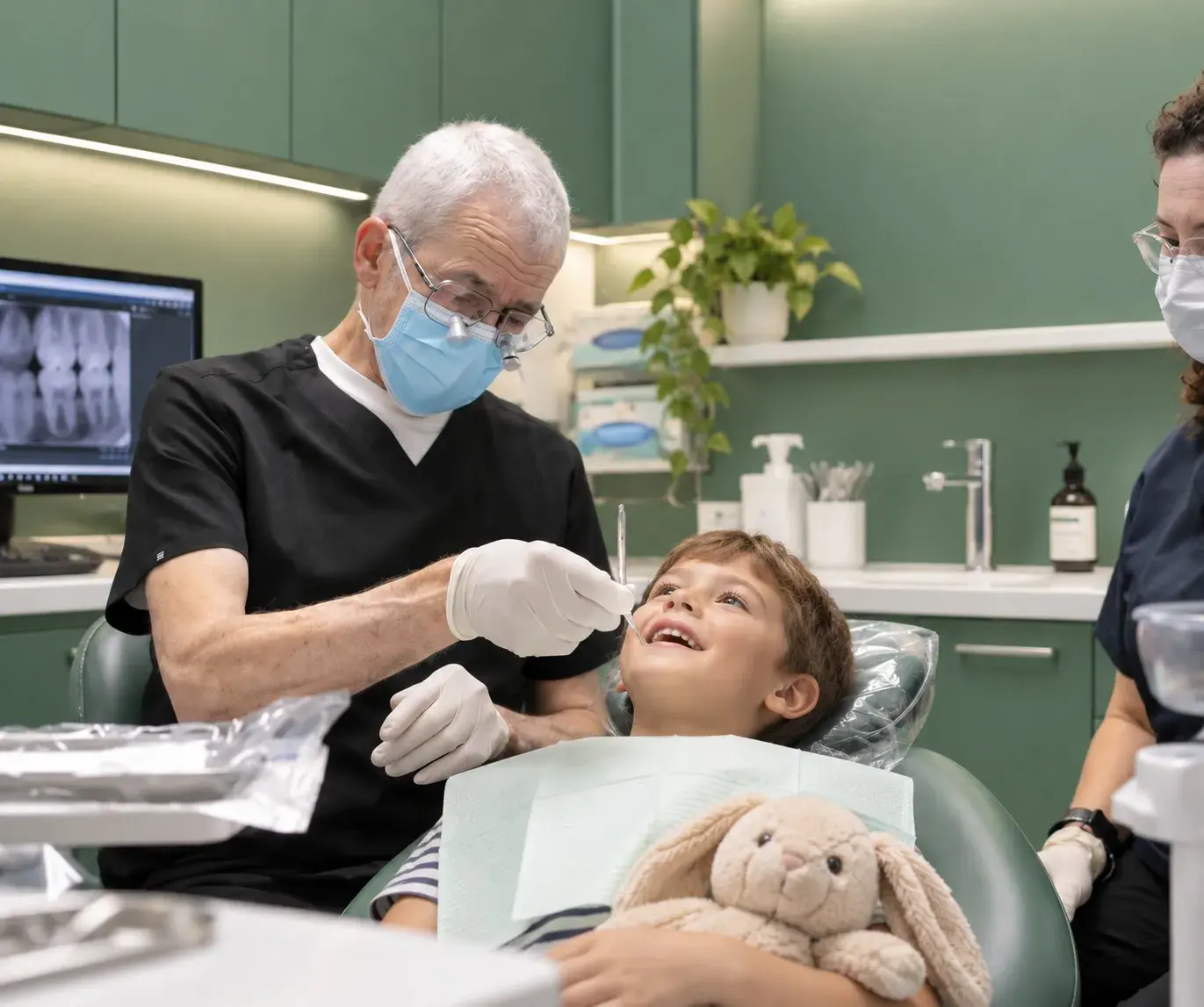 A dentist checking a smiling young boy's teeth as he hugs a toy bunny in the chair, his X-rays on the screen behind