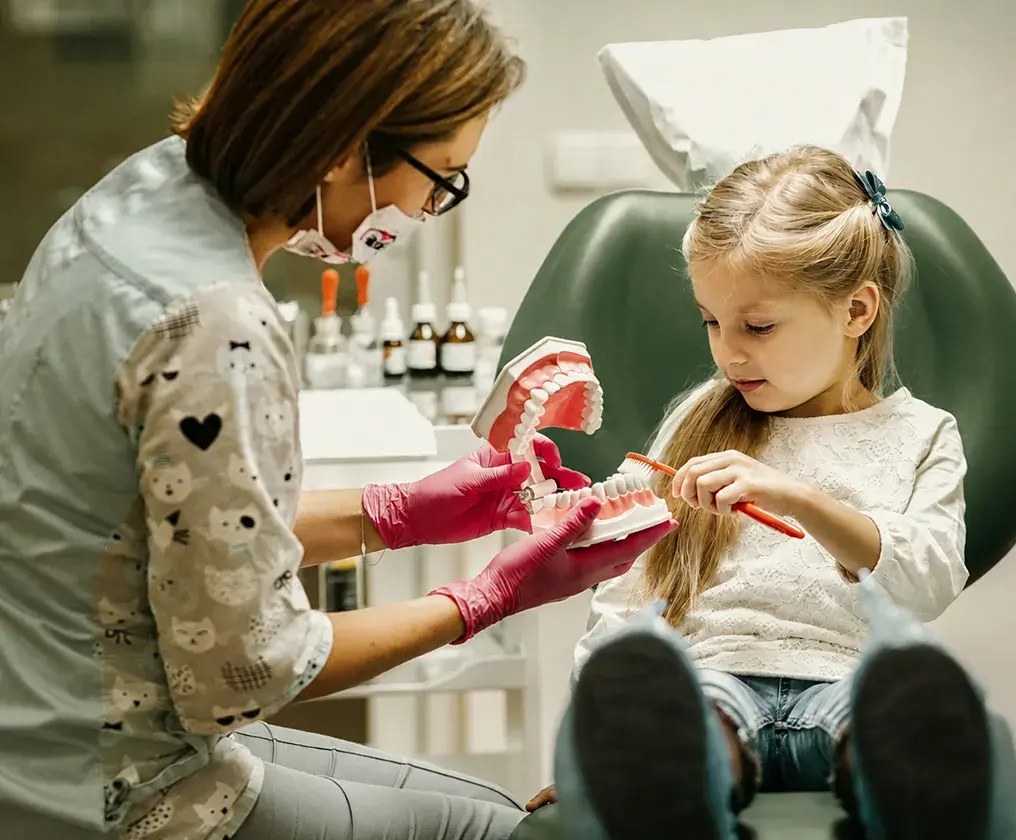 A dental clinician showing a young girl how to brush, using a large model of teeth while she holds a toothbrush