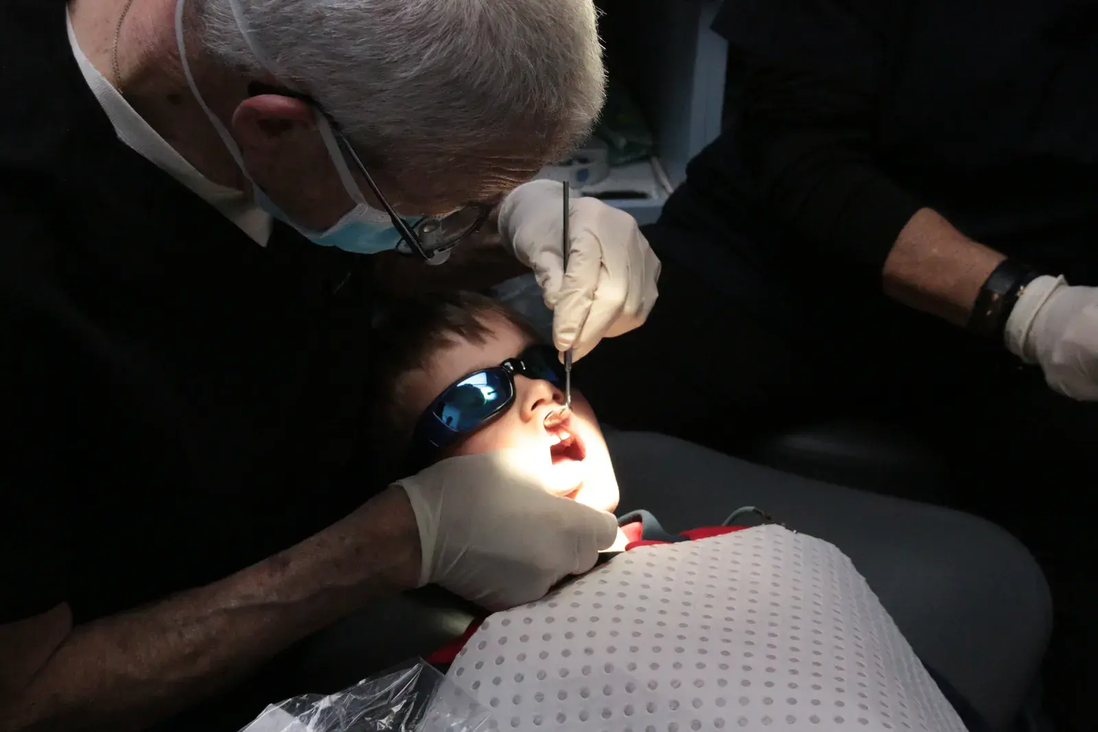 A dentist working carefully on a young patient's teeth under the light, the child wearing blue protective glasses