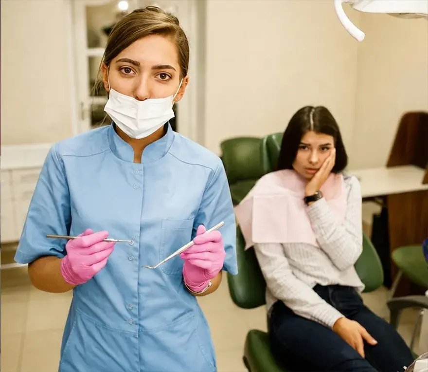 A dental assistant holding instruments, with a patient seated in the treatment chair behind her